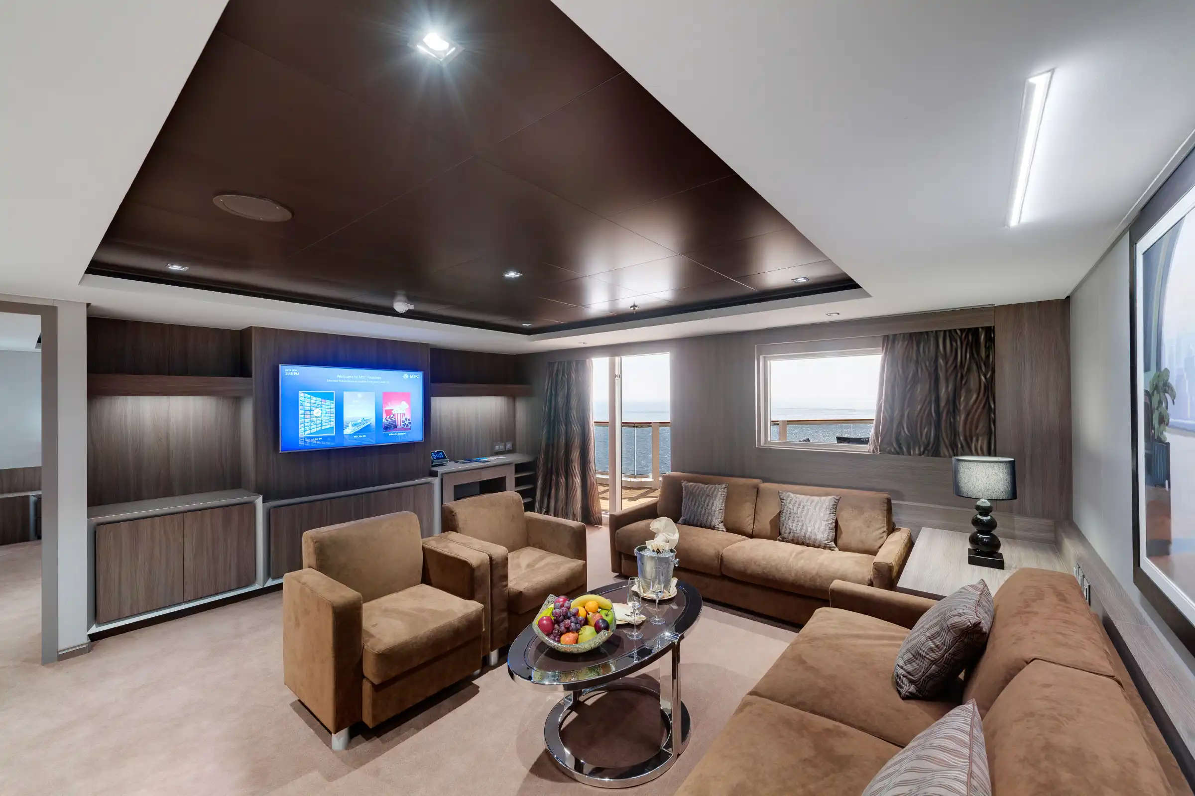 An upscale cruise ship suite featuring a spacious living area with multiple seating zones including a sofa, armchairs, and a round table with refreshments. The room showcases contemporary luxury design with dark wood ceiling details, panoramic windows with ocean views, integrated entertainment system, and warm neutral furnishings.