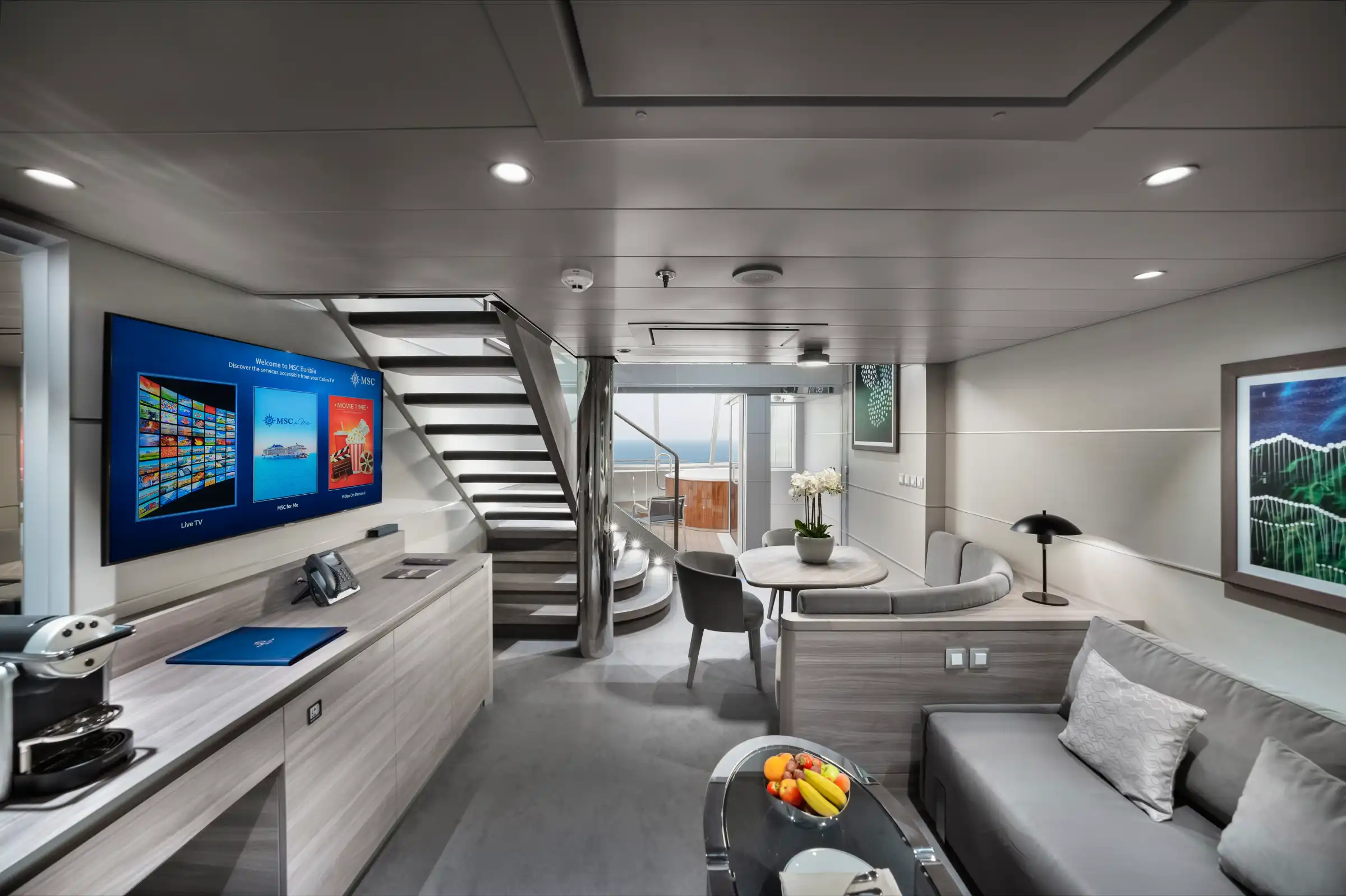 A modern two-level luxury cruise ship suite featuring a split-level layout with living area below, sleeping quarters above accessed by central staircase, integrated smart TV, kitchenette, contemporary furnishings, and floor-to-ceiling windows with balcony access.