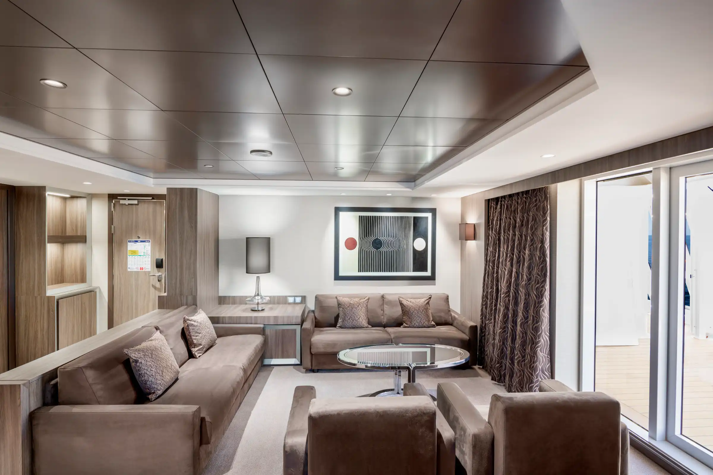 A luxurious cruise ship suite living area featuring dual taupe sofas arranged in an L-shape, contemporary furnishings, a metallic coffered ceiling with recessed lighting, floor-to-ceiling terrace doors, and elegant drapery. The space showcases premium finishes with wood paneling, modern artwork, and refined décor typical of high-end ship accommodations.