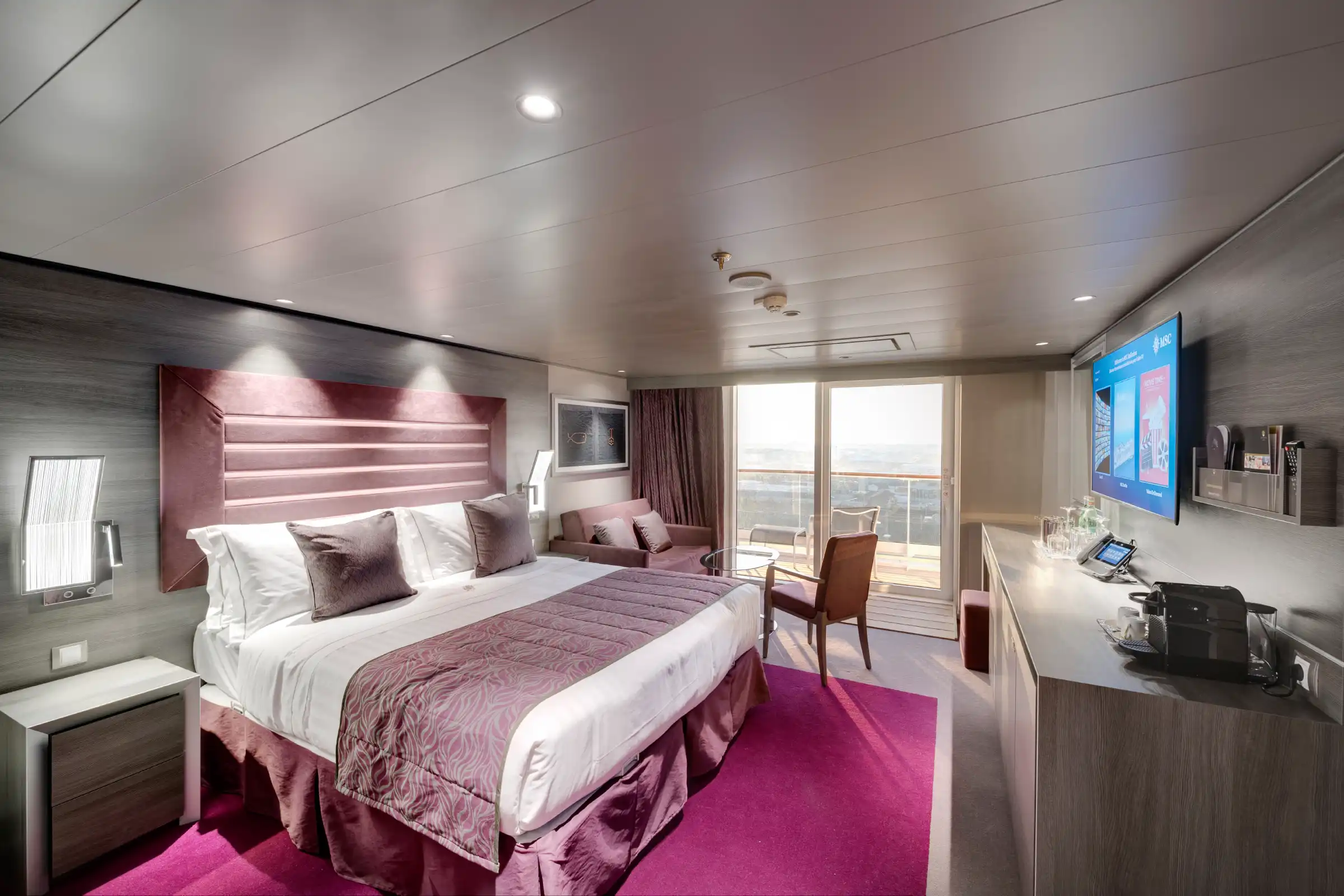 A luxury cruise ship suite featuring a king bed with burgundy upholstered headboard, white linens, and decorative pillows. The spacious cabin includes a seating area with armchair, flat-screen television, modern wood finishes, magenta accent rug, and large oceanview balcony with natural daylight.