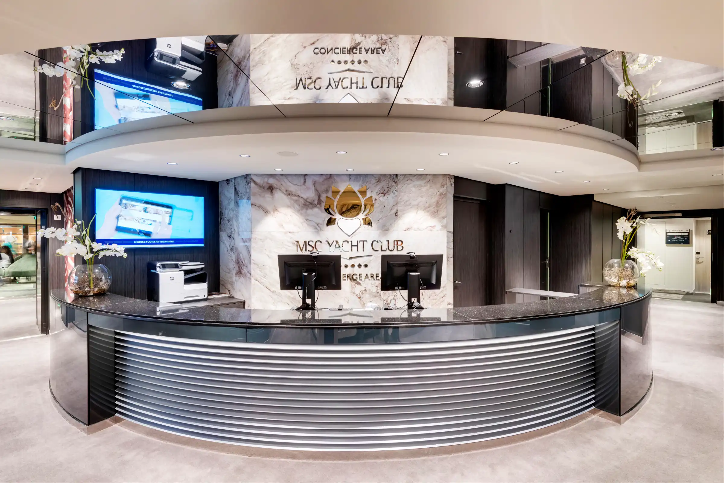 A modern, upscale concierge desk for an exclusive yacht club area, featuring curved metallic ribbed front panels in chrome/silver, white marble with gold emblem centerpiece, and two desktop workstations. The space is brightly lit with contemporary design elements including a curved ceiling and marble cladding, conveying premium service and luxury accommodation amenities.