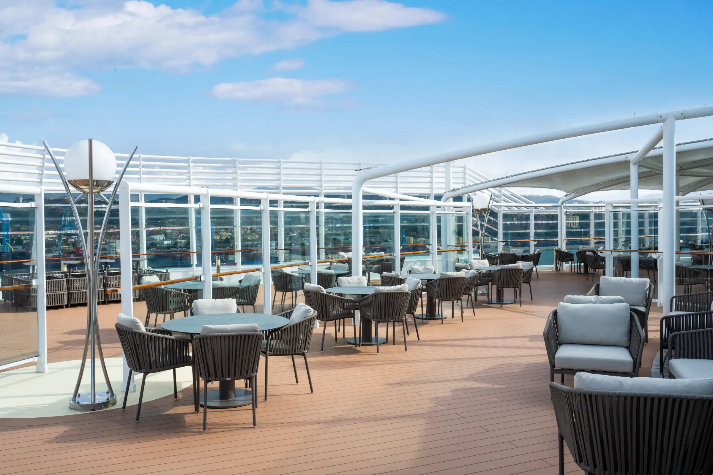 A spacious, modern rooftop bar venue with floor-to-ceiling windows offering panoramic sea views. The space features contemporary black rope-design chairs, round tables, and warm wood decking beneath a white structural frame, bathed in bright daylight.