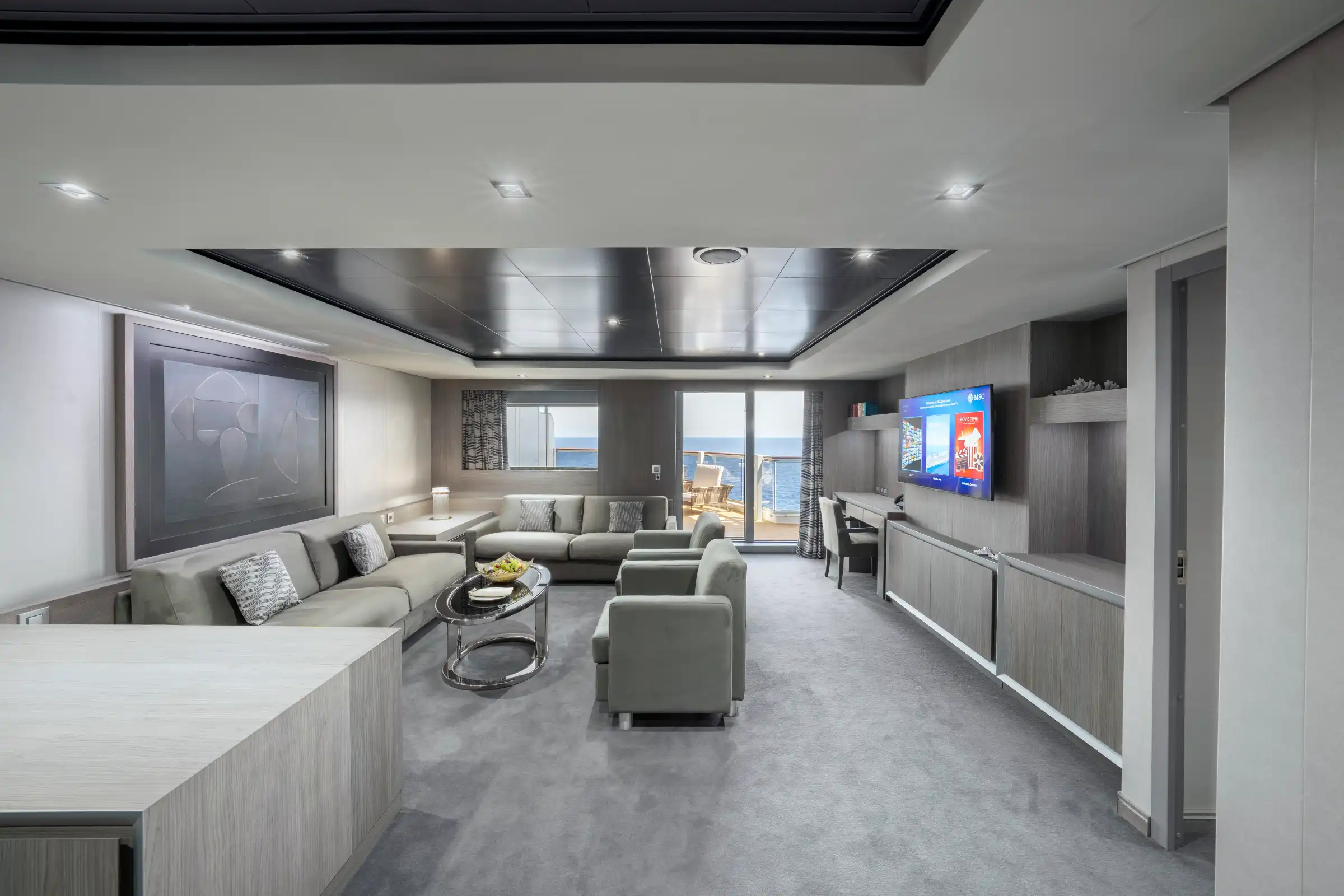 A luxury cruise ship suite featuring an expansive living area with a sectional sofa, modern furnishings, and a flat-screen TV mounted on the right wall. Large windows and a terrace door provide ocean views, while contemporary design elements include a metallic ceiling panel, recessed lighting, and high-end finishes throughout.