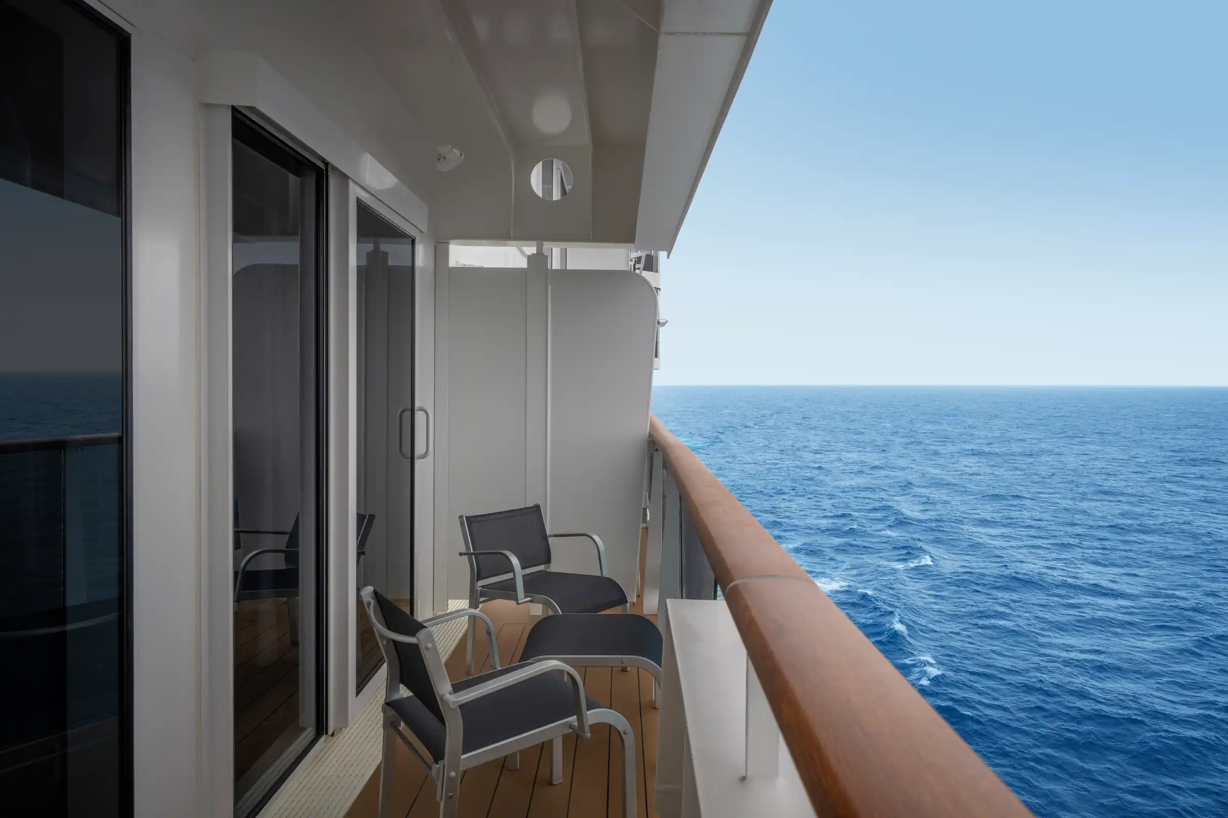 A private balcony of a luxury cruise ship suite featuring modern sliding glass doors leading to an outdoor seating area with two lounge chairs. The expansive ocean and clear sky dominate the background, showcasing the elevated vantage point and open-air amenities of the accommodation.
