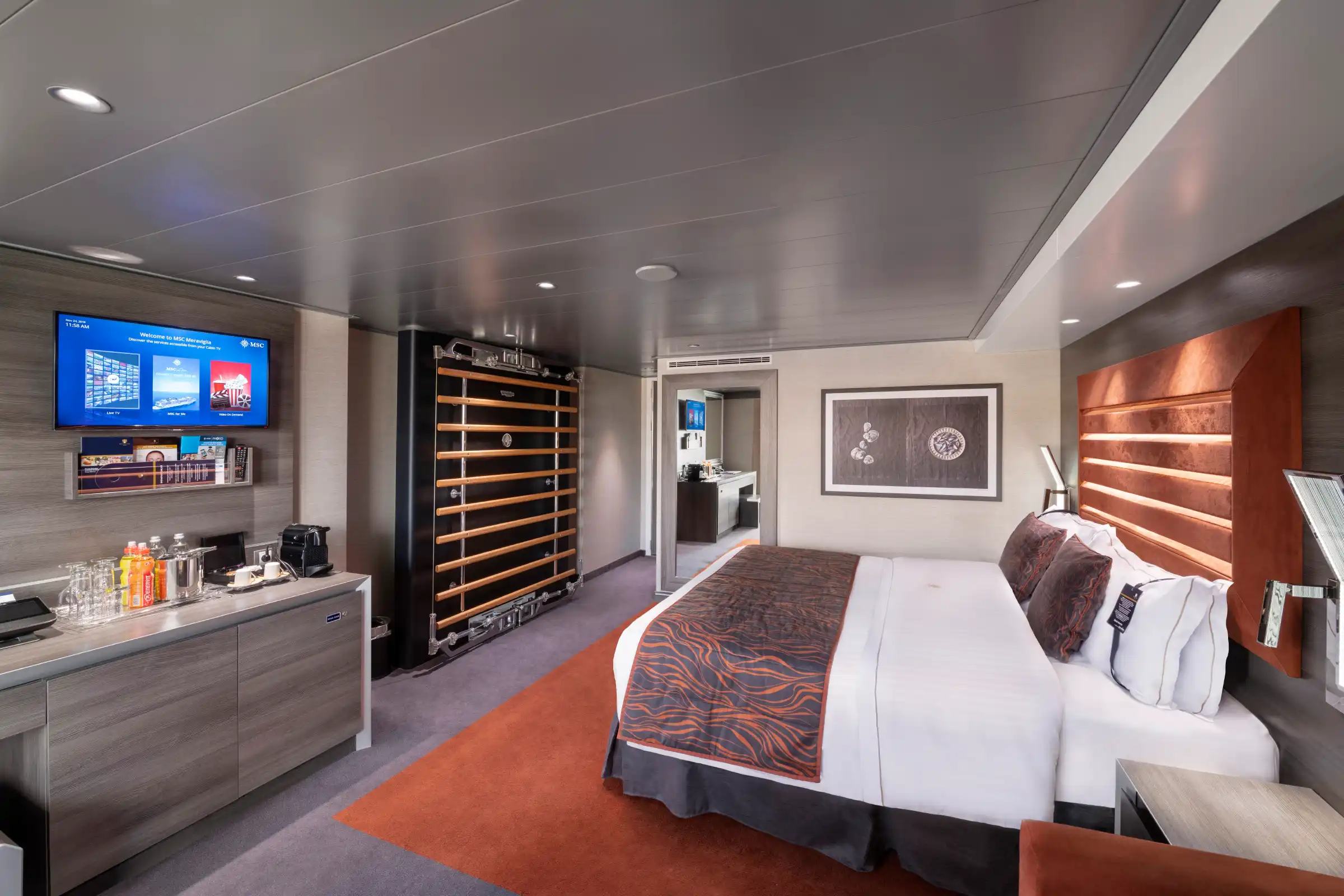 A luxury cruise ship stateroom featuring a queen bed with burgundy patterned bedding, wood-accent headboard with orange lighting, flat-screen TV mounted on gray feature wall, wine cooler, and contemporary furnishings. The suite displays modern maritime luxury design with warm accent lighting and neutral tones.