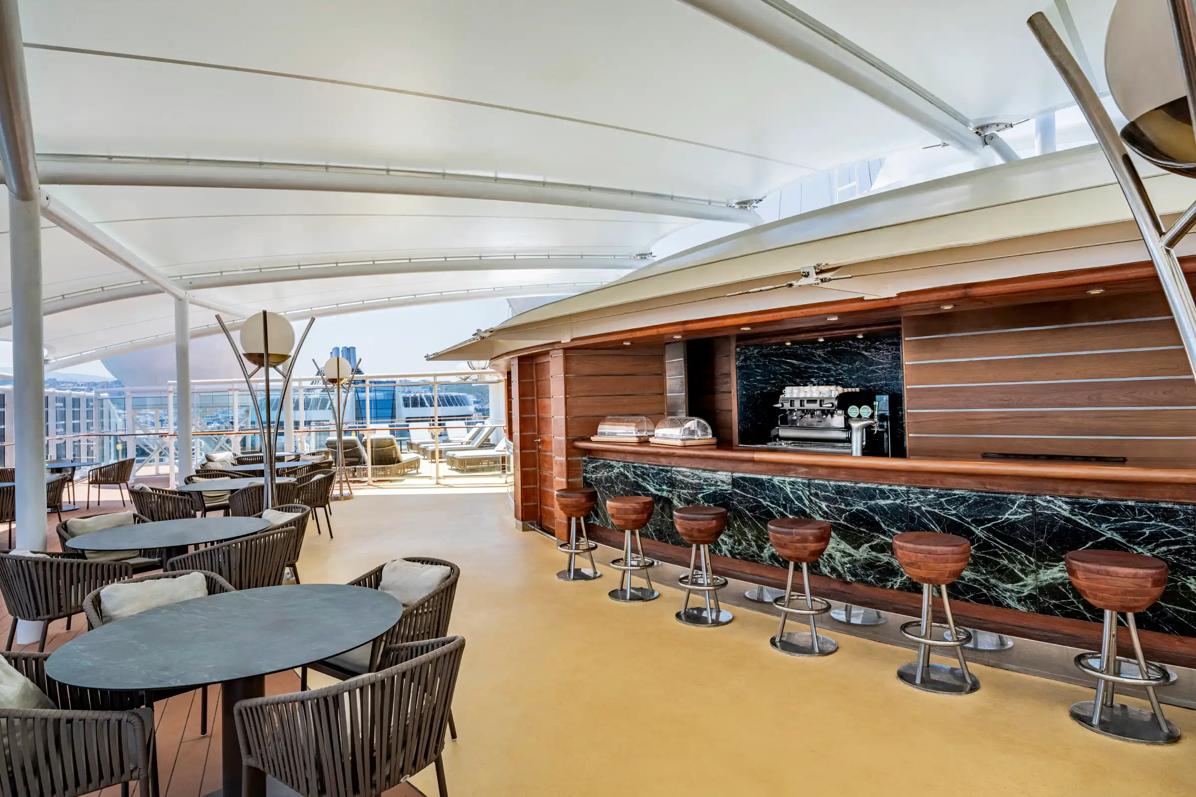 An upscale cruise ship bar lounge featuring a modern interior with a long bar counter backed by dark green marble, wooden seating areas with woven chairs, and an open retractable ceiling system. The venue offers both indoor and outdoor deck access with panoramic ocean views.