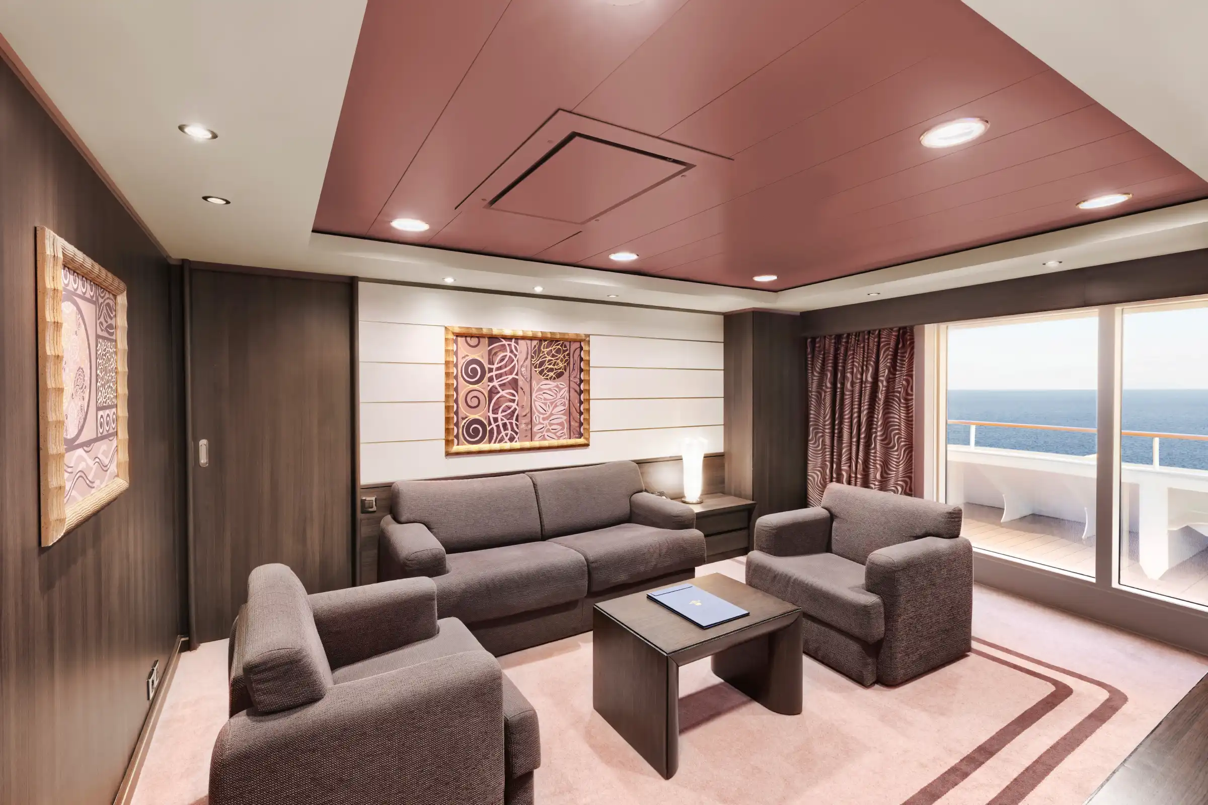 A luxury cruise ship suite with contemporary furnishings featuring grey upholstered seating, dark wood paneling, and a distinctive rose-tinted coffered ceiling. Large glass doors open to a balcony overlooking the ocean, with decorative framed artwork and recessed LED lighting throughout.