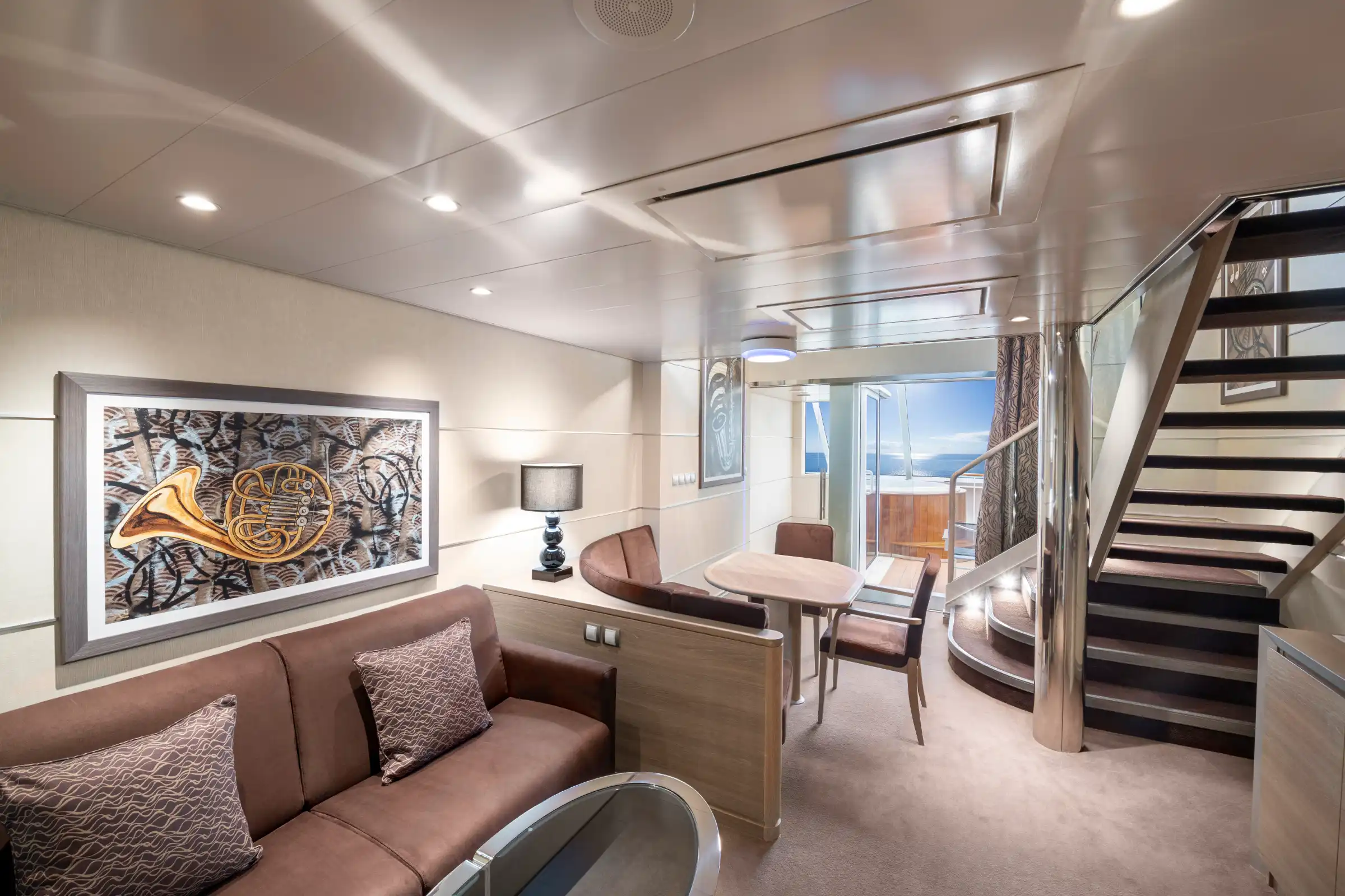 A luxurious two-level cruise ship suite featuring a spacious lower level with contemporary furnishings including a brown sofa, dining area, framed artwork, and access to a private balcony with ocean views. An elegant staircase leads to an upper level, with modern lighting and high-end finishes throughout.