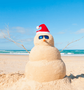 Schneemann aus Sand mit Sonnenbrille und rot-weißer Weihnachtsmütze am Strand bei blauem Himmel.