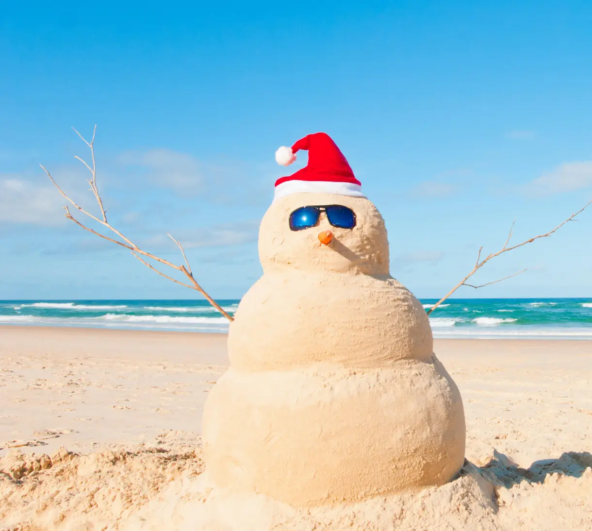 Schneemann aus Sand mit Sonnenbrille und rot-weißer Weihnachtsmütze am Strand bei blauem Himmel.