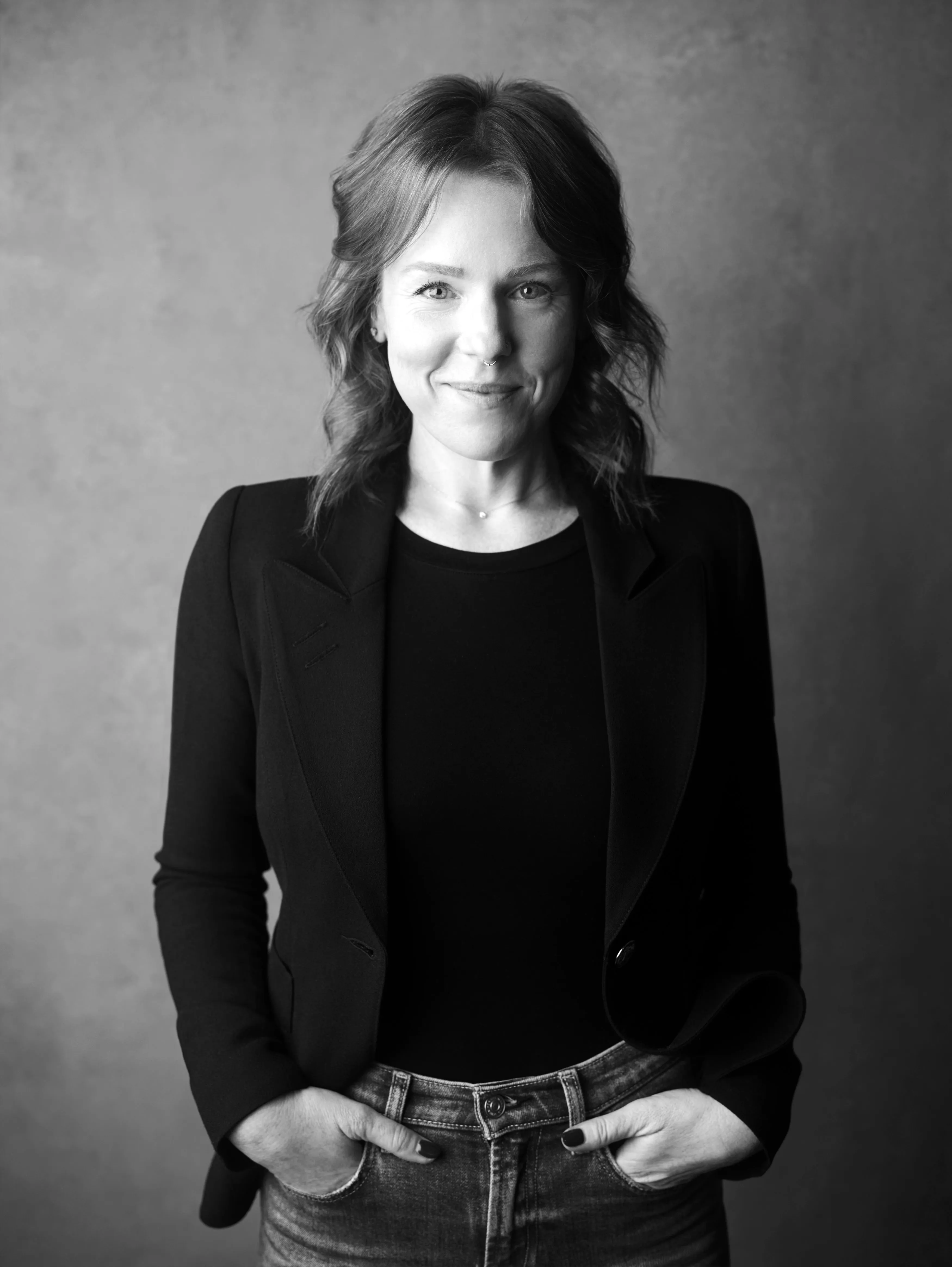 Smiling woman with wavy hair in a black blazer and jeans, hands in pockets, against a plain background.