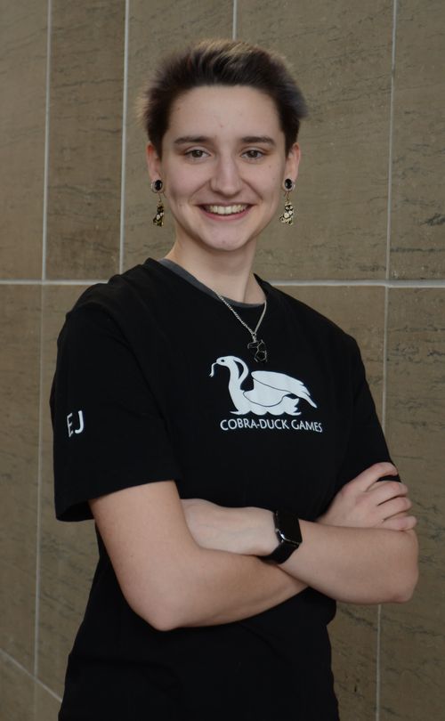 This is a portrait of Elis in a black t-shirt with the Cobra Duck Games logo on it. They are smiling with their arms crossed.