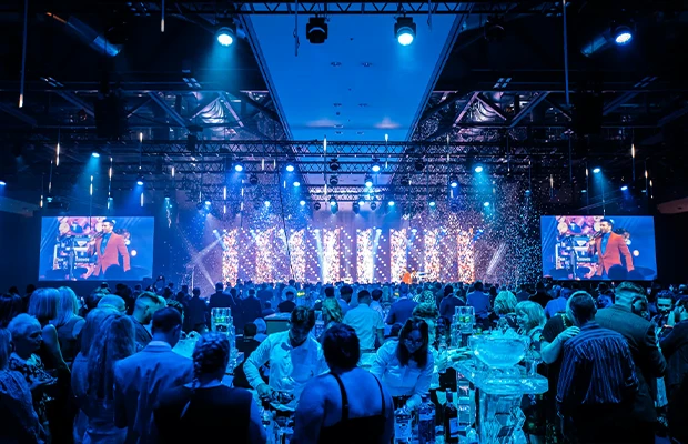 A photo of a large indoor event with people, decorative lighting, and screens showing a speaker.