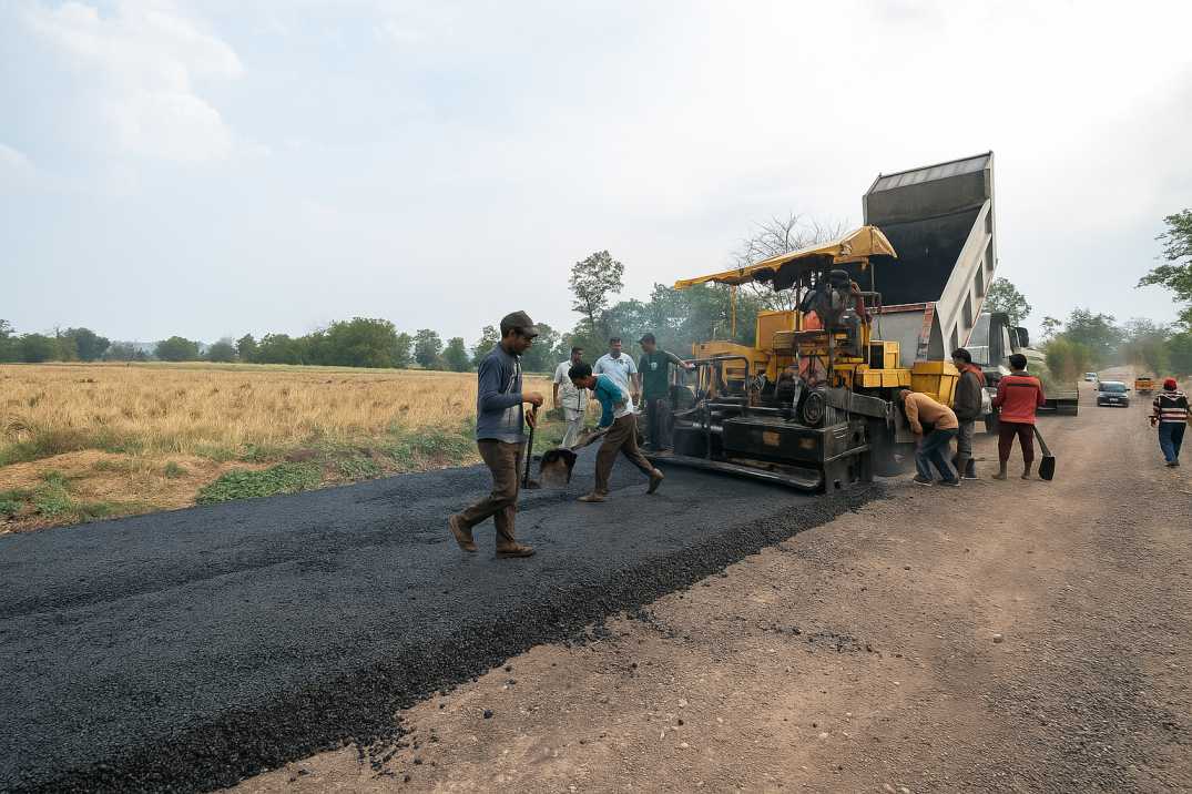 Workers laying fresh asphalt on a rural road using a yellow paver and dump truck.