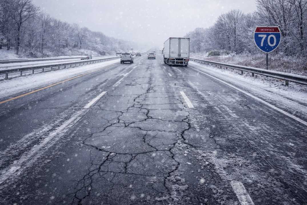 Snow-covered U.S. highway with icy surface and visible pavement cracking under overcast winter sky.