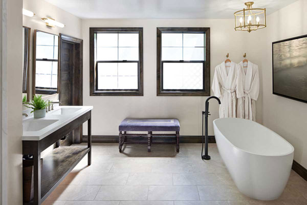 Modern bathroom with freestanding white bathtub, dark wood-framed windows, double sink vanity, purple upholstered bench, and two white robes hanging on wall hooks.