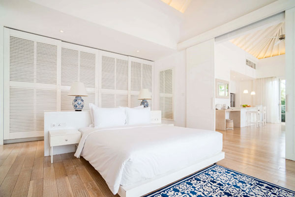 Bright spacious bedroom with white bed linens, wooden floor, blue patterned rug, and lamps on bedside tables.