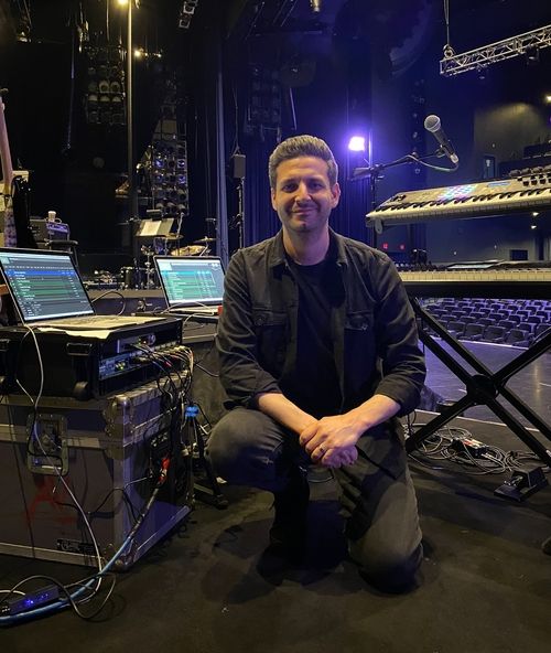 Gabe Rudner kneels in front of his keyboards and other musical equipment on stage in a large music venue