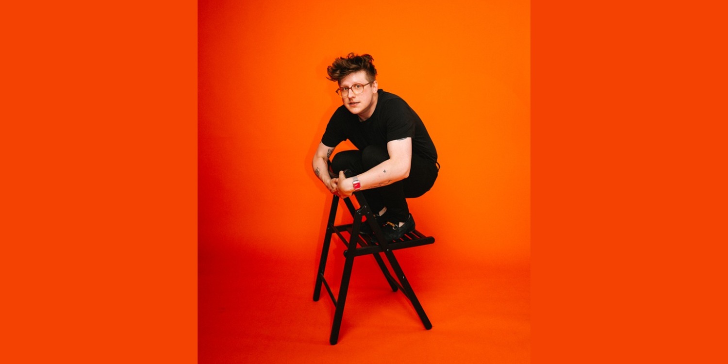 Hinnerk Köhn hockt auf einem Klappstuhl vor orangenen Hintergrund.