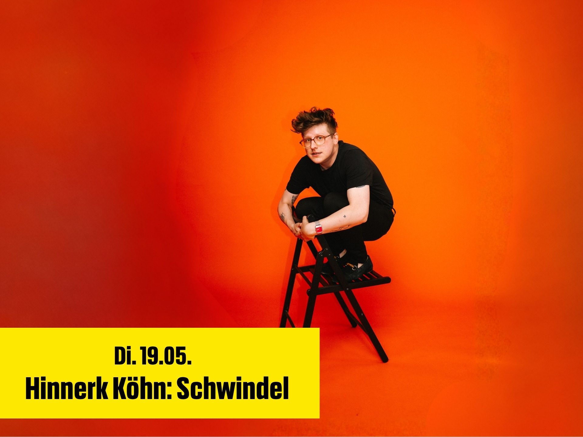 Der Comedian Hinnerk Köhn sitz falsch herum auf einem schwarzen Stuhl vor einem roten Hintergrund