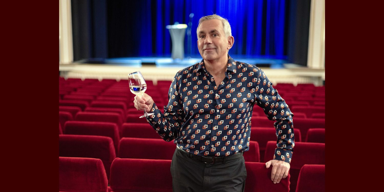 Kaminer in einem Theatersaal zwischen den Stühlen stehend mit Weinglas in der Hand.