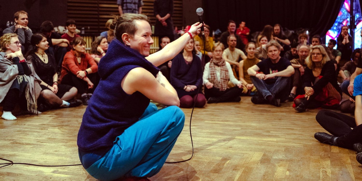 Die Workshopleiterin Öyven Jonatta hockt in der Mitte eines Personenkreises und erklärt etwas. Um sie herum sitzen die Teilnehmer.