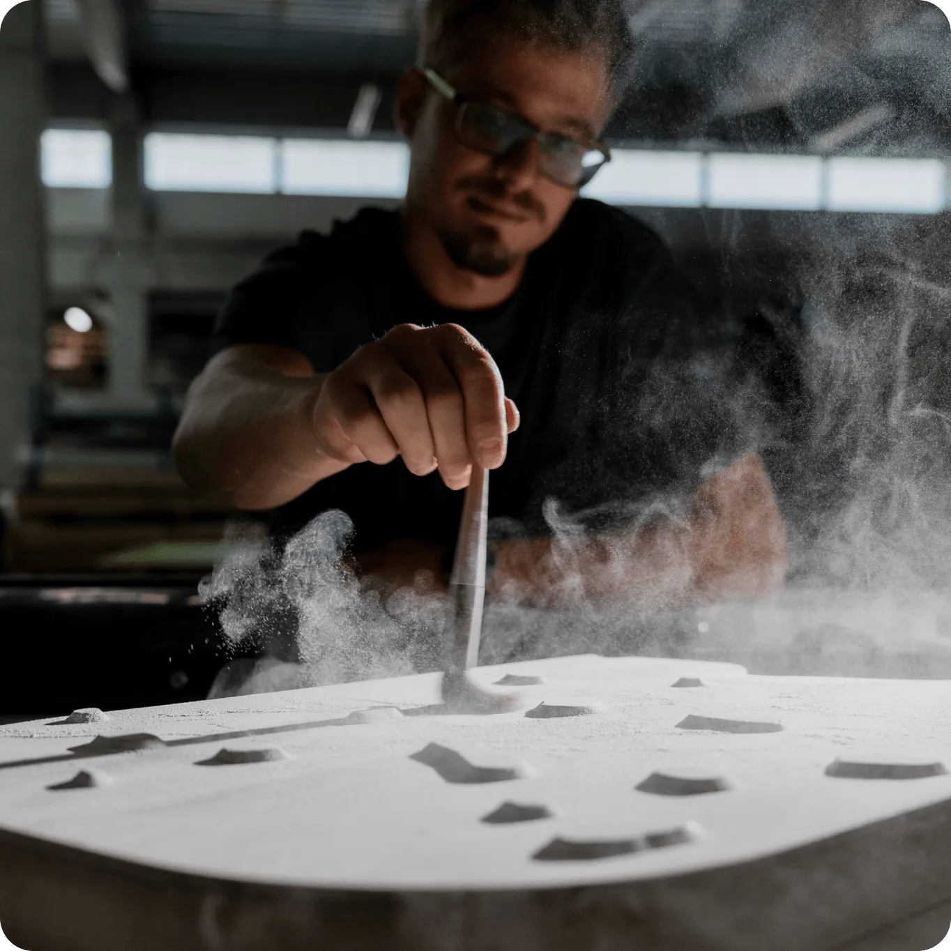 Muž s brýlemi ručně čistí nebo modeluje bílý práškový materiál ve výrobním prostředí.