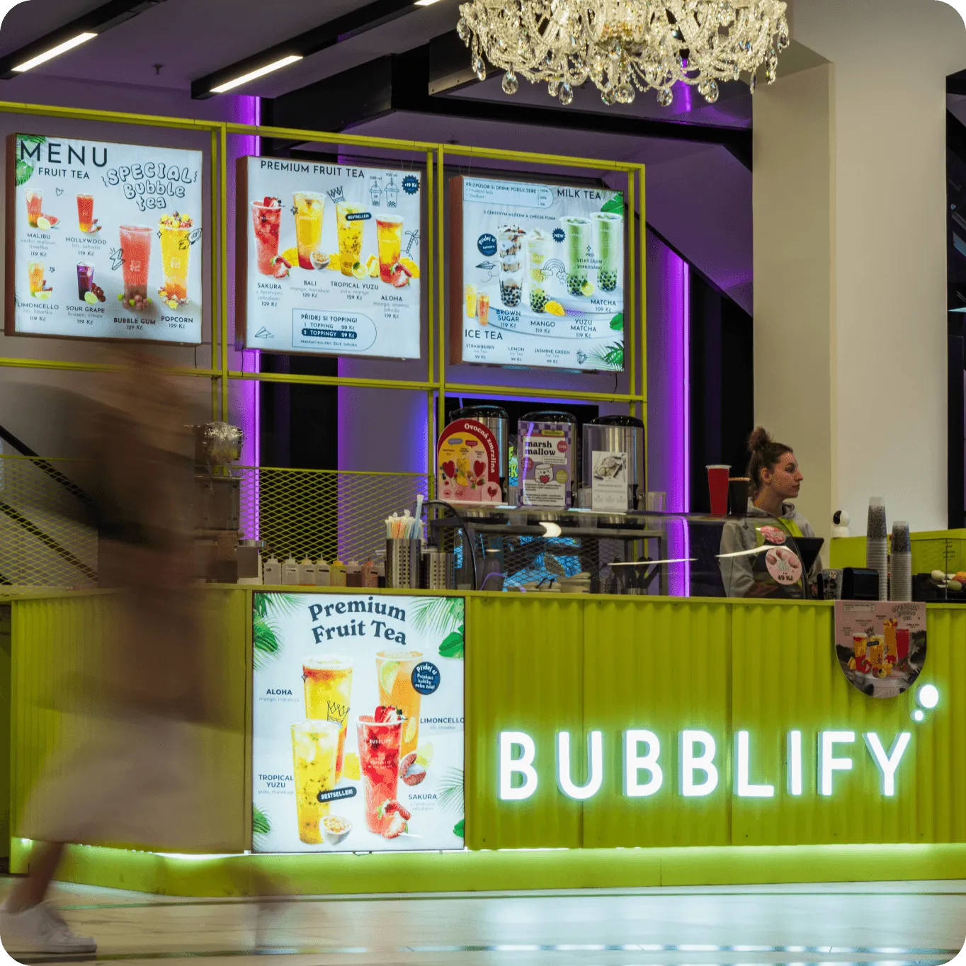 Stánek BUBBLIFY s nabídkou ovocných a mléčných čajů, fialové osvětlení a krystalový lustr.