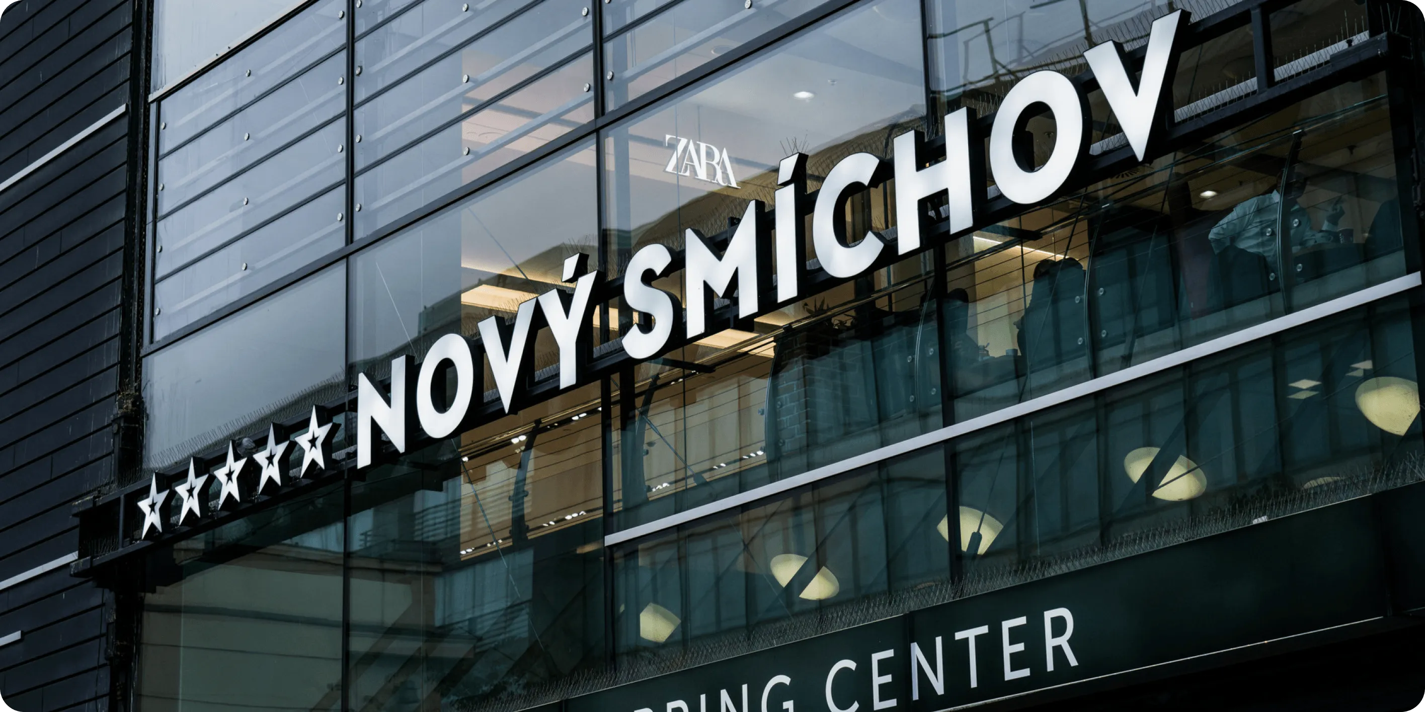 Nápis Nový Smíchov na prosklené fasádě nákupního centra s odrazem okolních budov.