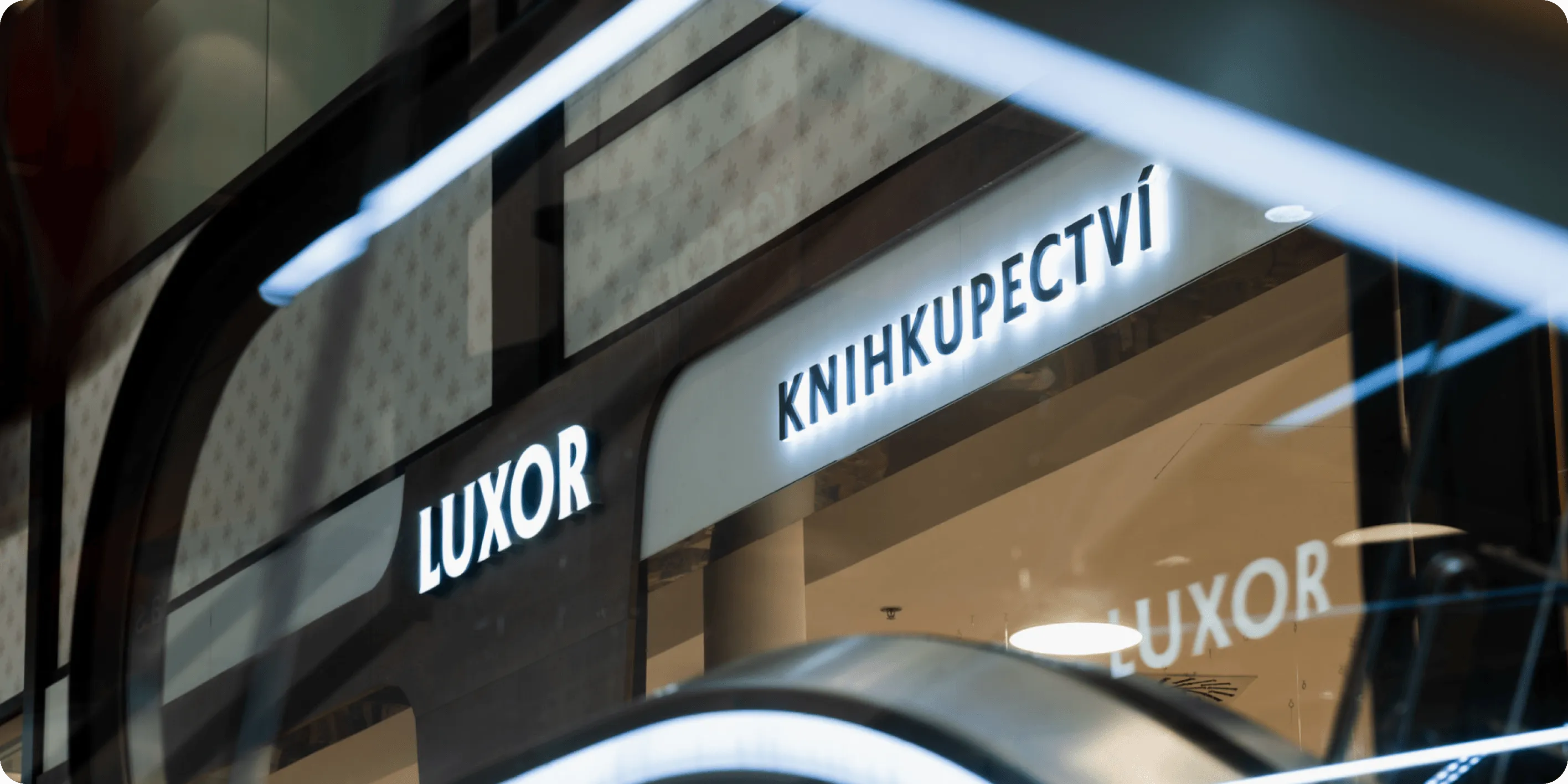 Svítící nápisy LUXOR a KNIHKUPECTVÍ na fasádě knihkupectví viděné skrz prosklenou plochu.