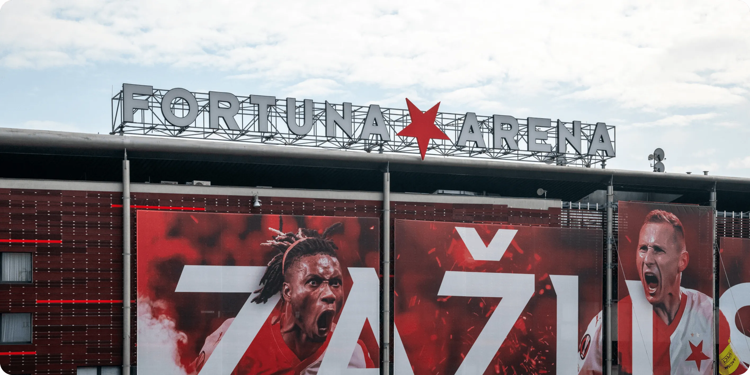 Exteriér stadionu Fortuna Areny s velkými plakáty dvou křičících fotbalistů v červené a bílé.