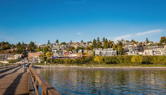 Waterfront residential neighborhood with houses on a hillside and people walking on a wooden pier under clear blue sky.