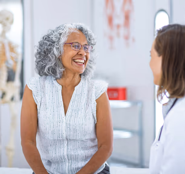 Smiling elderly woman with gray curly hair and glasses talking to a doctor in a medical office.