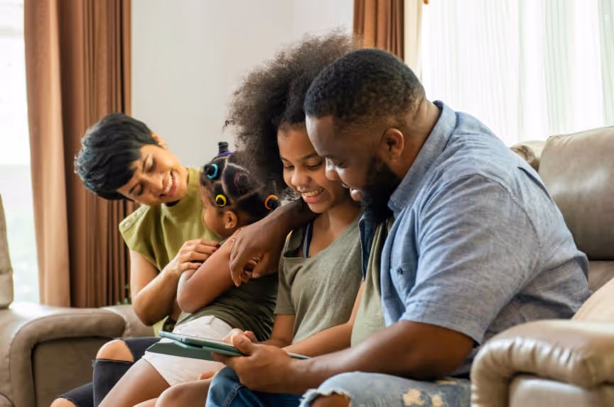 Smiling family of four sitting on a couch, looking at a tablet together.