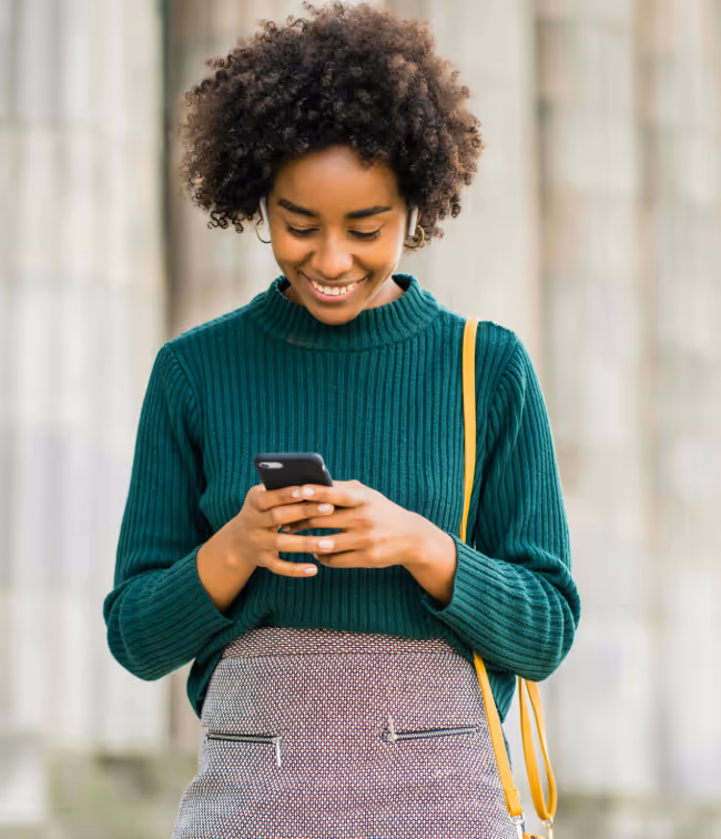 Smiling woman with curly hair in a green sweater looking at her phone while standing outdoors.