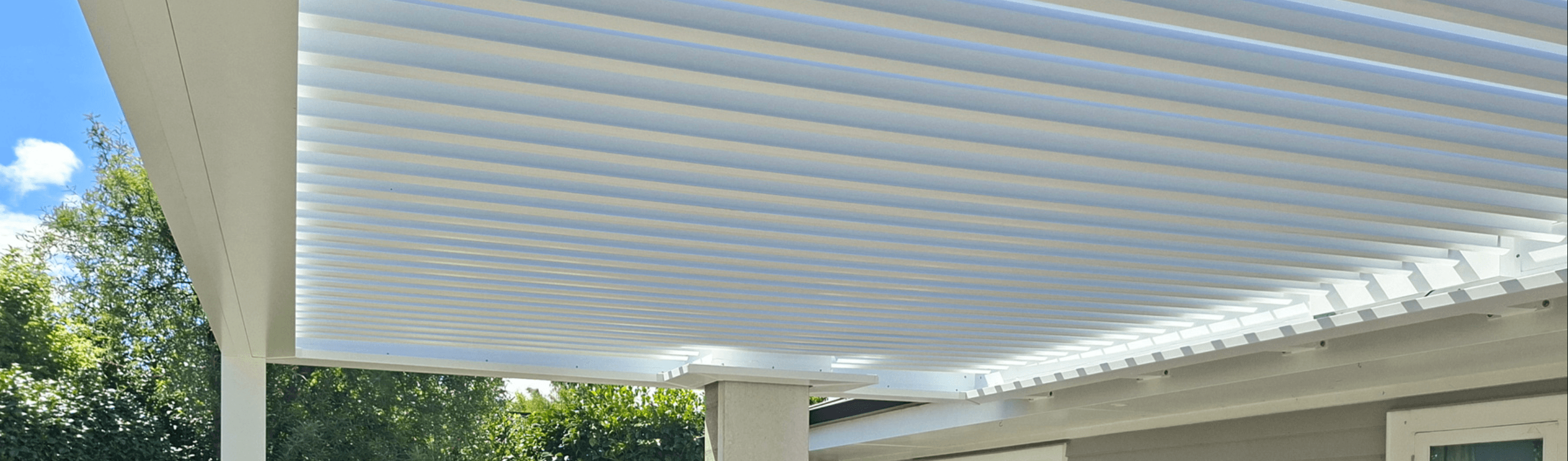 White pergola roof with adjustable slats above outdoor space with trees in the background.
