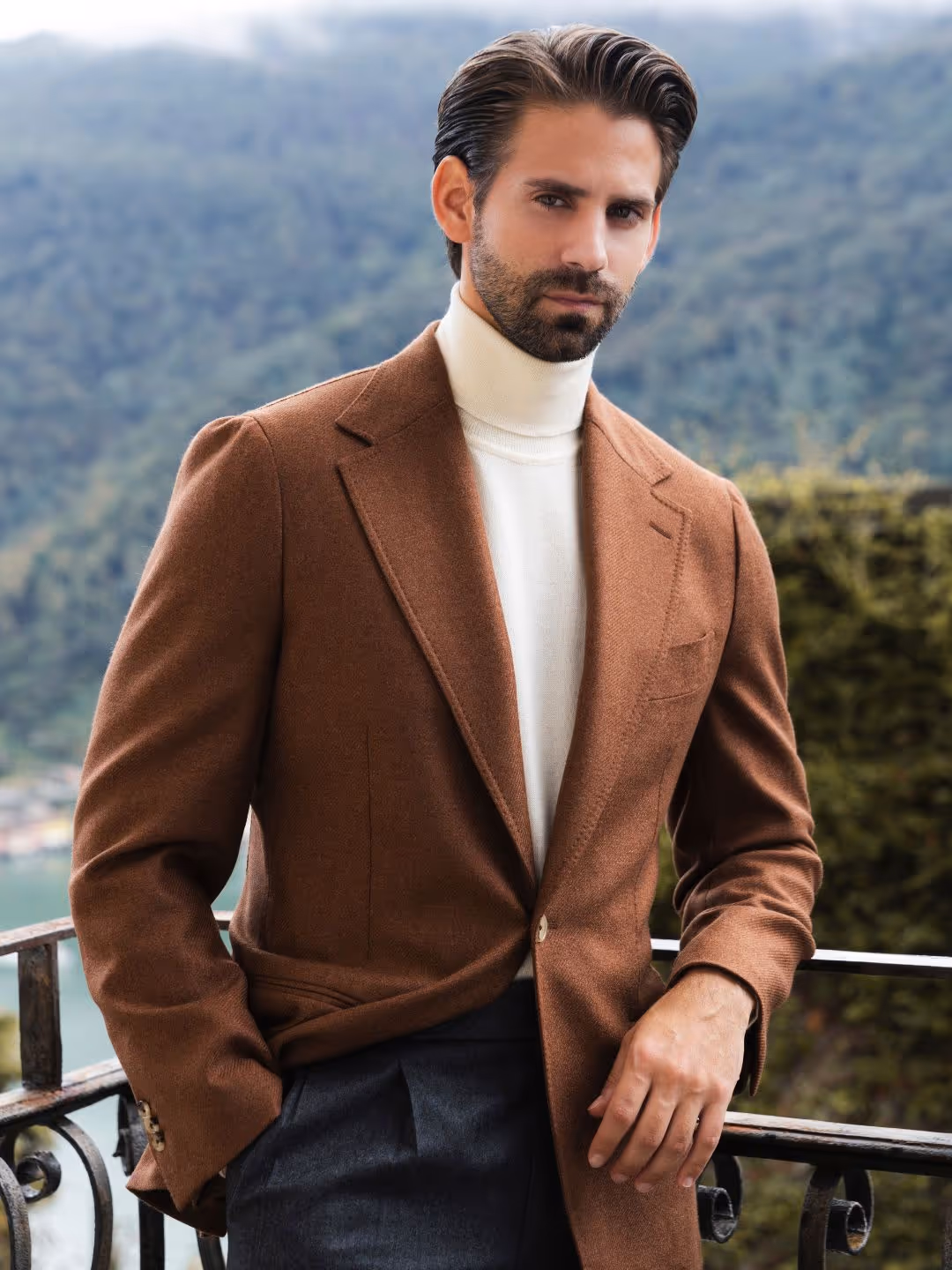 Mann mit braunem Mantel und weißem Rollkragenpullover lehnt an einem Balkon mit Wald und Hügeln im Hintergrund.