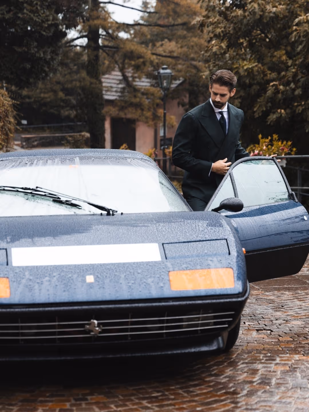 Mann im Anzug öffnet Tür eines blauen Oldtimer-Sportwagens bei regnerischem Wetter.