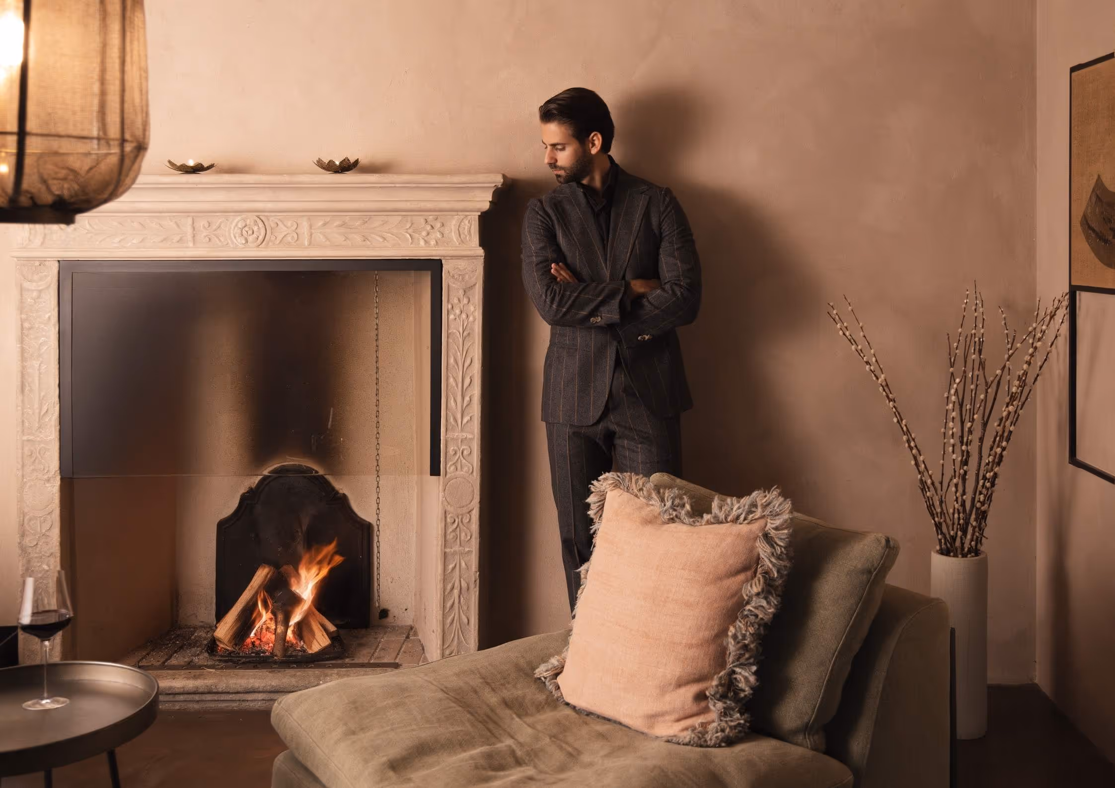 Ein Mann im dunkel karierten Anzug lehnt mit verschränkten Armen an einer Wand neben einem Kamin mit brennendem Holzfeuer in einem warm eingerichteten Wohnzimmer.