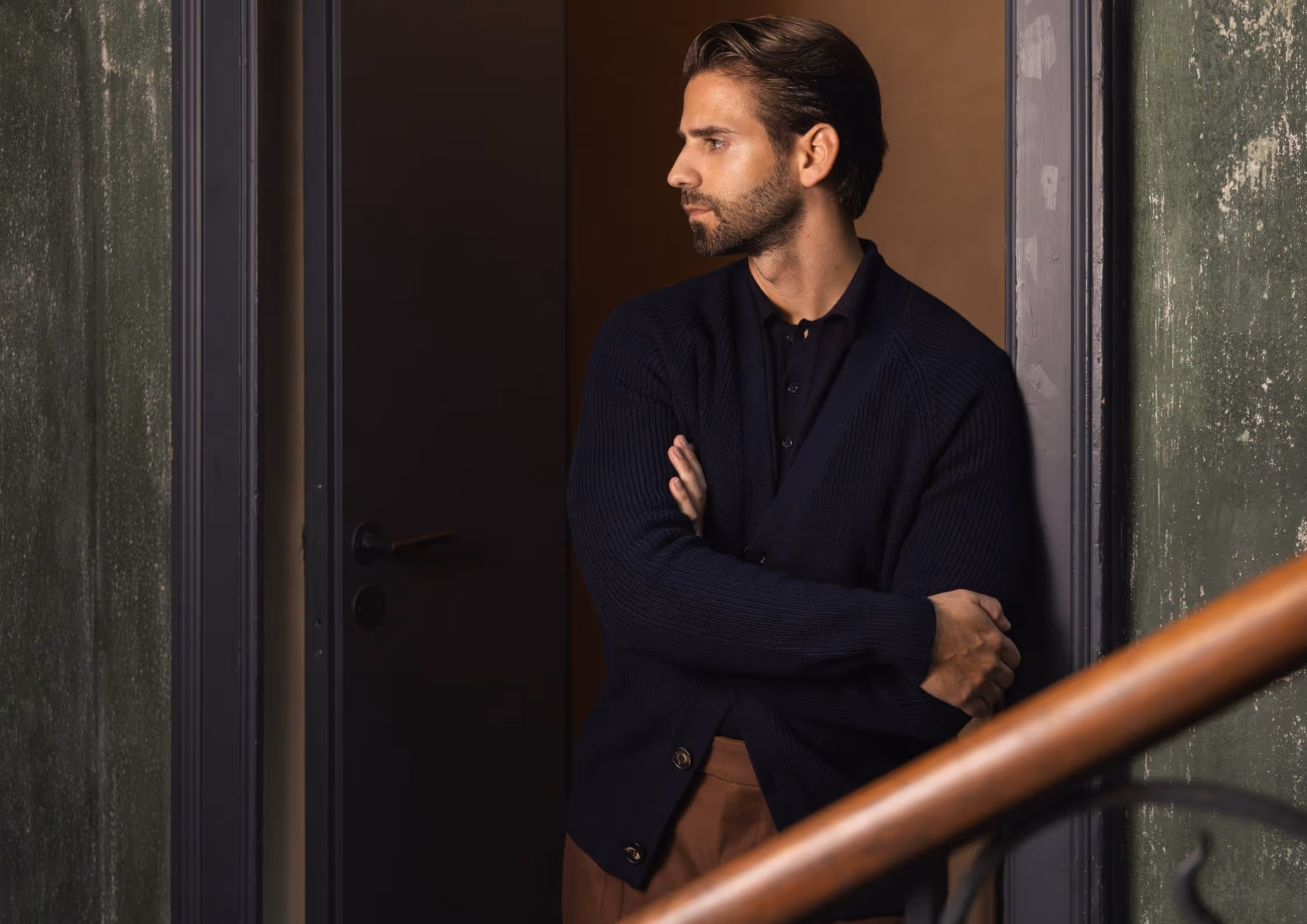 Ein Mann mit dunklem Pullover lehnt an einem Türrahmen und blickt nach rechts.