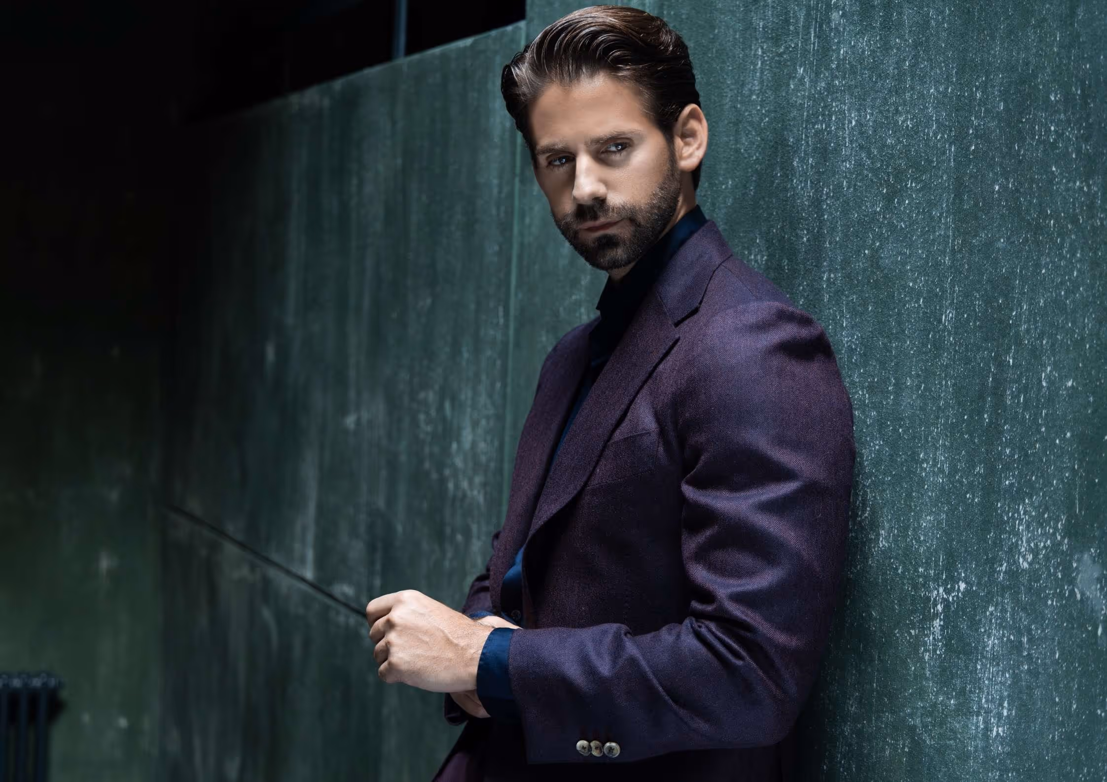 Ein Mann mit braunem Haar und Bart lehnt sich an eine grüne Wand, trägt ein dunkelviolettes Sakko und blickt ernst.