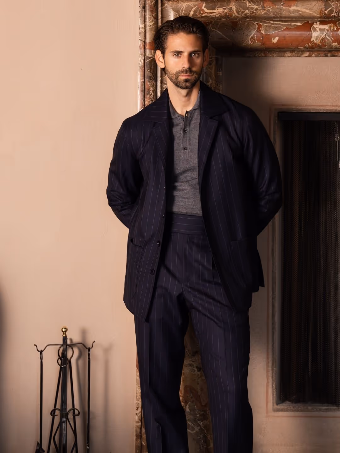 Mann mit Bart und dunklem, gestreiftem Anzug lehnt vor einem Kamin mit hellbeiger Wand im Hintergrund.