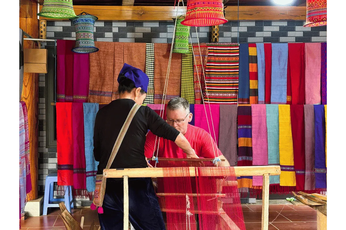 An ethnic women helping a tourist to weave a traditional fabric on a wooden loom, surrounded by colorful textiles in Pu Luong.