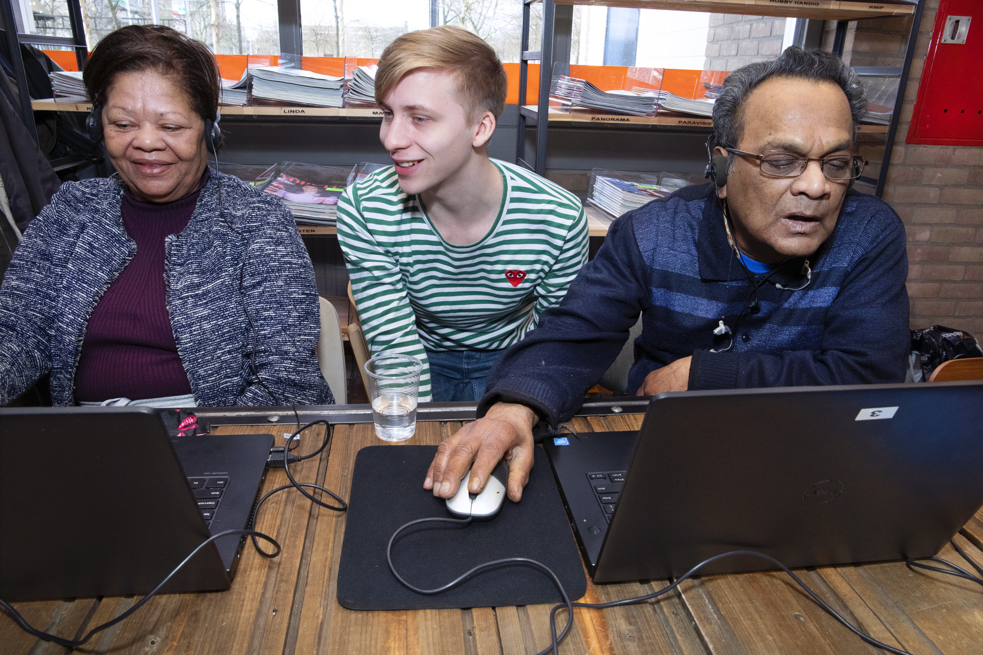Foto van een jonge, lachende MDT deelnemer die tussen twee oudere mensen aan tafel zit. De oudere mensen hebben elk een opengeklapte laptop voor zich, de jonge deelnemer kijkt mee met één van hen. 
