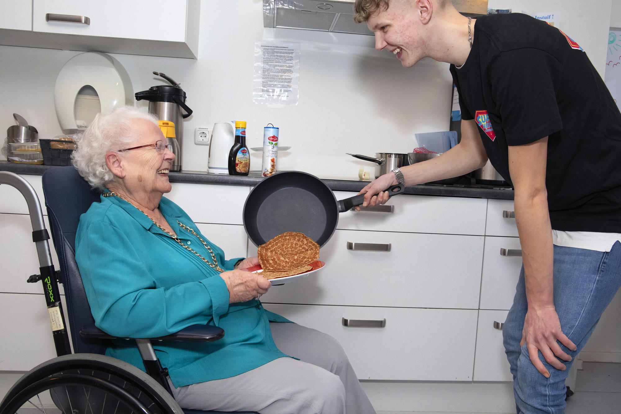 Foto van een jonge MDT deelnemer die een pannenkoek uitserveert aan een oudere vrouw in een rolstoel. 