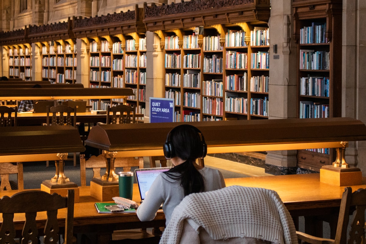 Student studying at a laptop in a university-style library with tall bookshelves