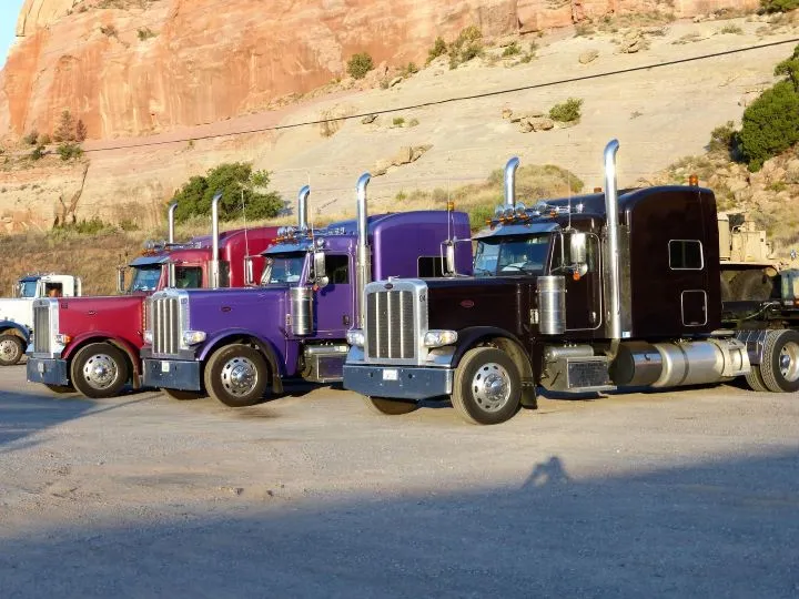 Several colorful semi-trucks, including red, purple, blue, and black vehicles, are parked along a street beside a tan stone wall.