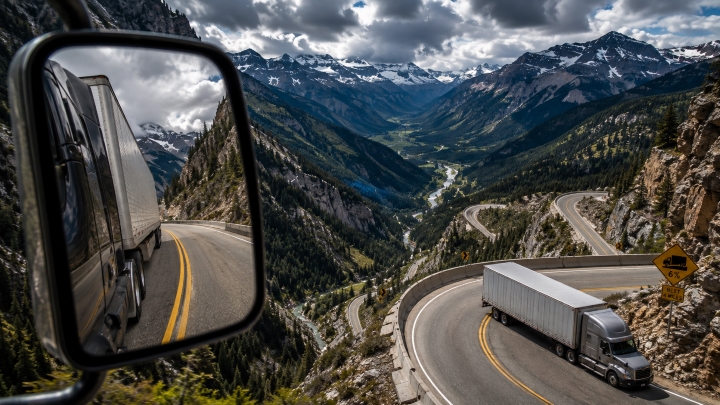 Semi-truck descending a winding mountain pass seen through its side mirror