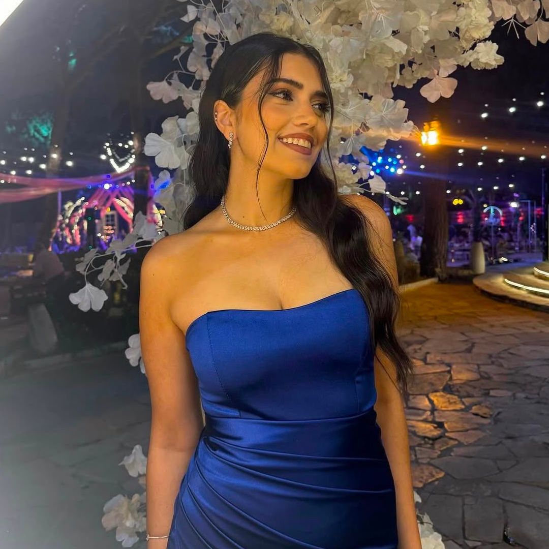Woman in a strapless blue satin dress smiles with white flowers and night lights in the background.