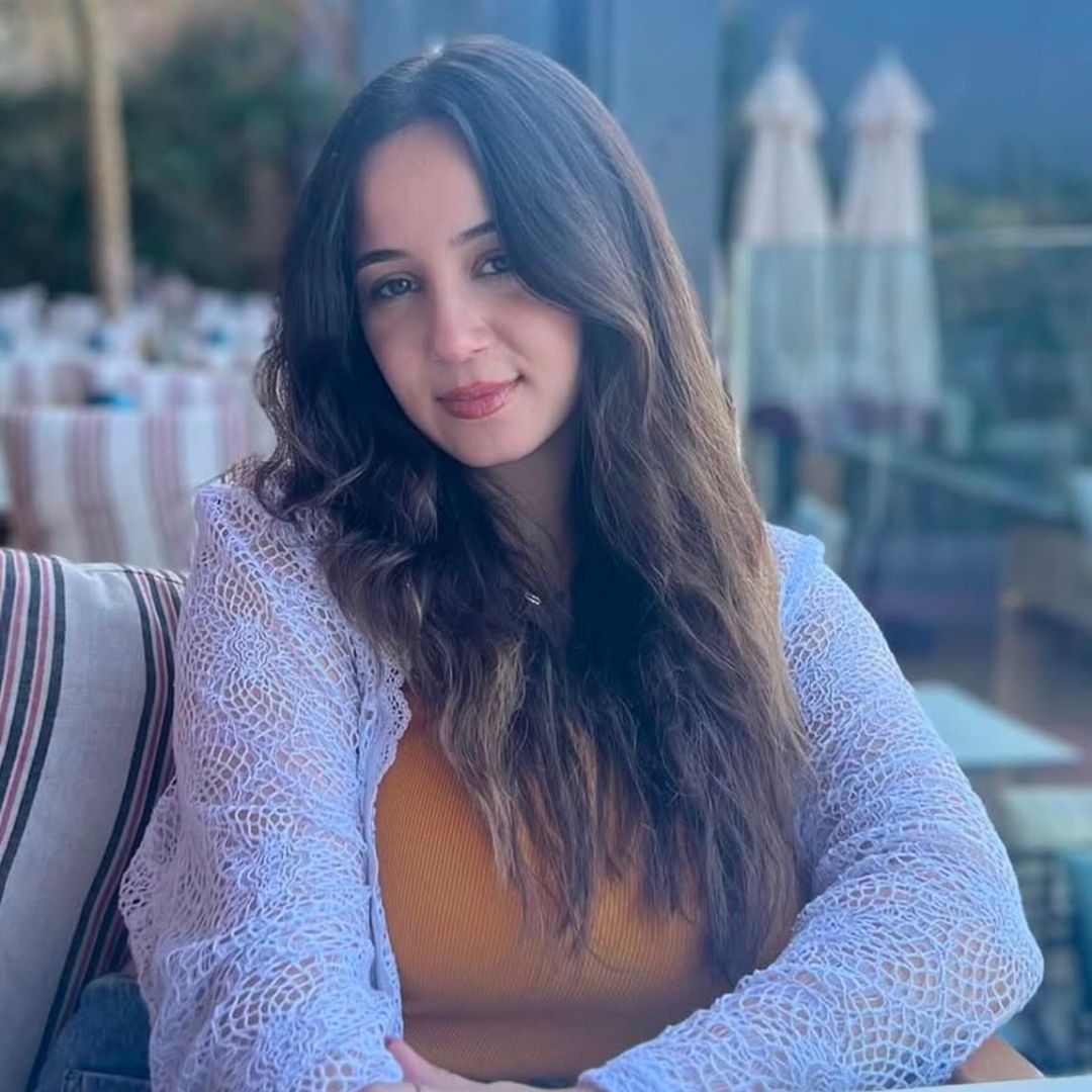 Young woman with long wavy hair wearing a white lace cardigan over a mustard top, sitting outdoors.